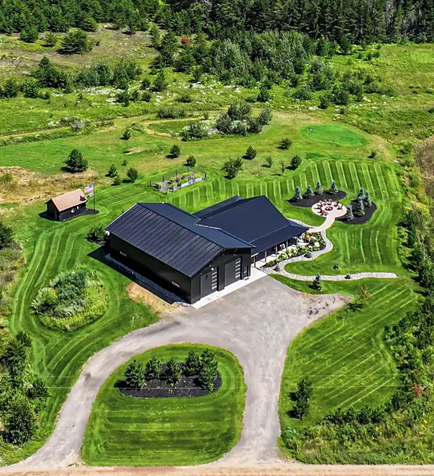 Aerial view of a black pole shop with striped mowed lawn, curving drive, planting islands and a paver fire pit circle