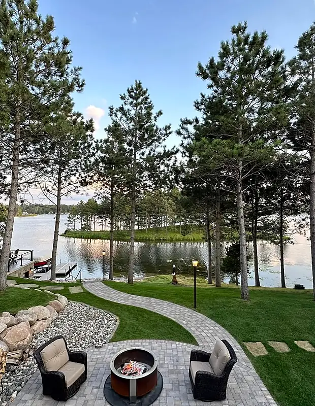 Paver fire pit patio with a curving walkway running down a pine-shaded lawn to a dock at dusk