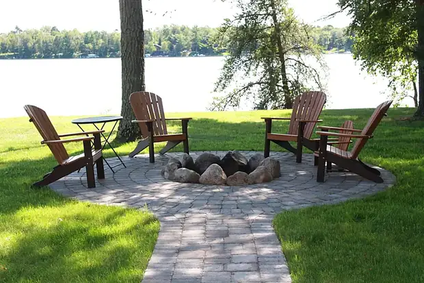 Circular paver patio with a boulder fire pit ring and Adirondack chairs overlooking a lake