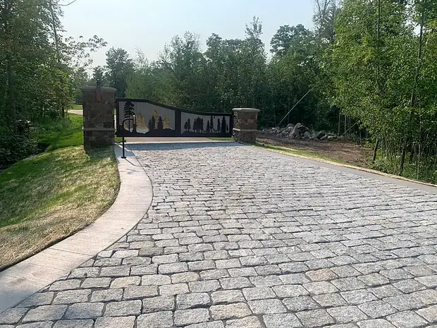 Wide cobblestone paver driveway curving toward a stone-pillared entry gate