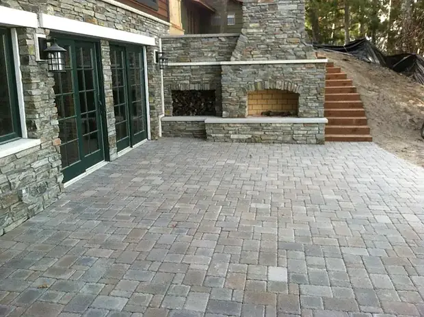 Large paver patio with a built-in stone outdoor fireplace and woodbox beside a walkout entry
