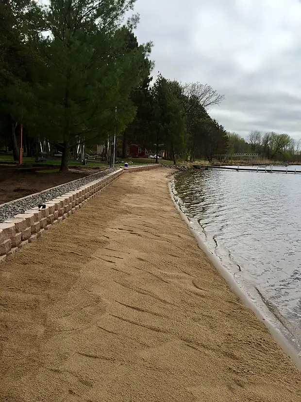 New sand beach along a lake edge held by a low block seawall with riprap beyond