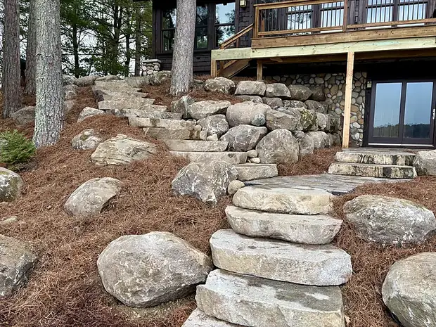 Wide natural stone slab steps descending a wooded slope between boulders to a lakeside home