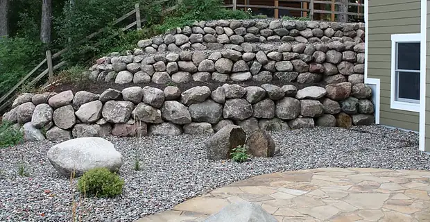 Three-tier natural boulder retaining wall stepping up a steep slope beside a home