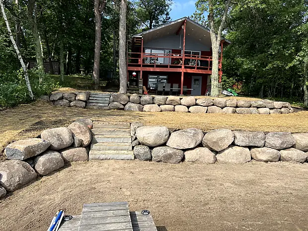 Two tiers of large boulder retaining wall stepping up a slope to a red lakeside cabin